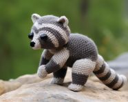 Raccoon crocheted in gray and grey and stands on a flat rock, crocheted style art